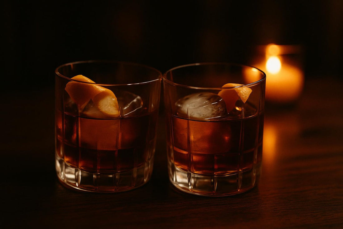 Two Old Fashioneds touching at the rim like a toast in candlelight — anniversary cocktail