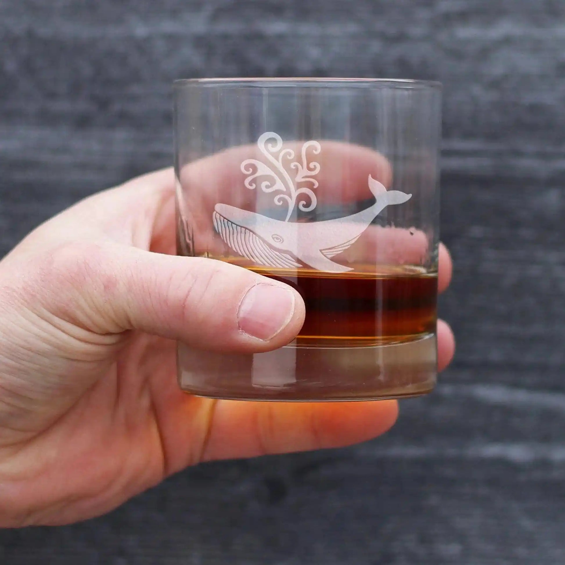Hand holding a glass with a whale design and liquid inside against a dark background